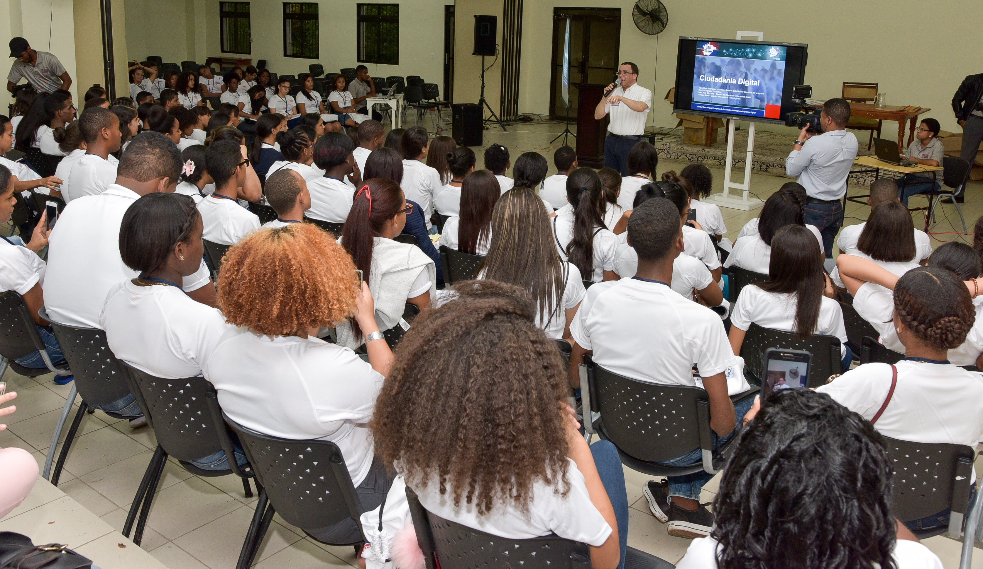  imagen Ministro Andrés Navarro de pie expone con microfono frete a estudiantes participantes del Foro Nacional Estudiantil los cuales se encuentran sentados escuchando atentamente. 
