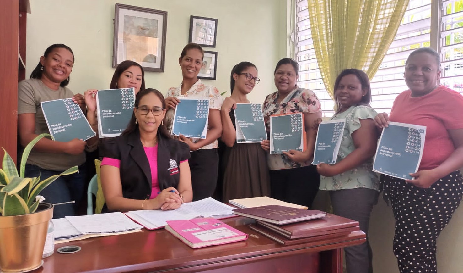 Imagen de una encargada sentada en escritorio, detrás un grupo de participantes con folletos en mano sobre Plan de Autodesarrollo personal